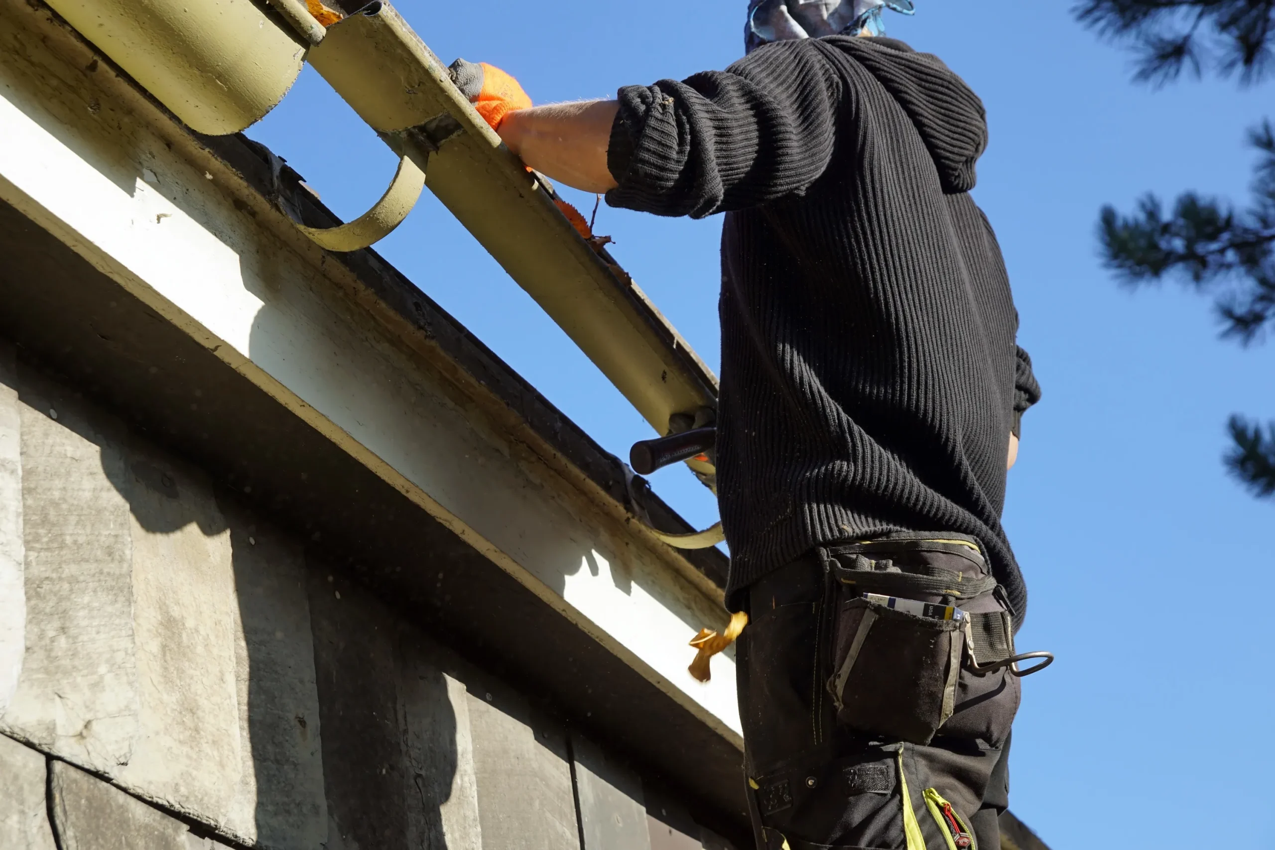 A worker in a black hoodie cleans a roof gutter on a sunny day. They wear orange gloves and stand on a ladder against the building. Tall trees are in the background.