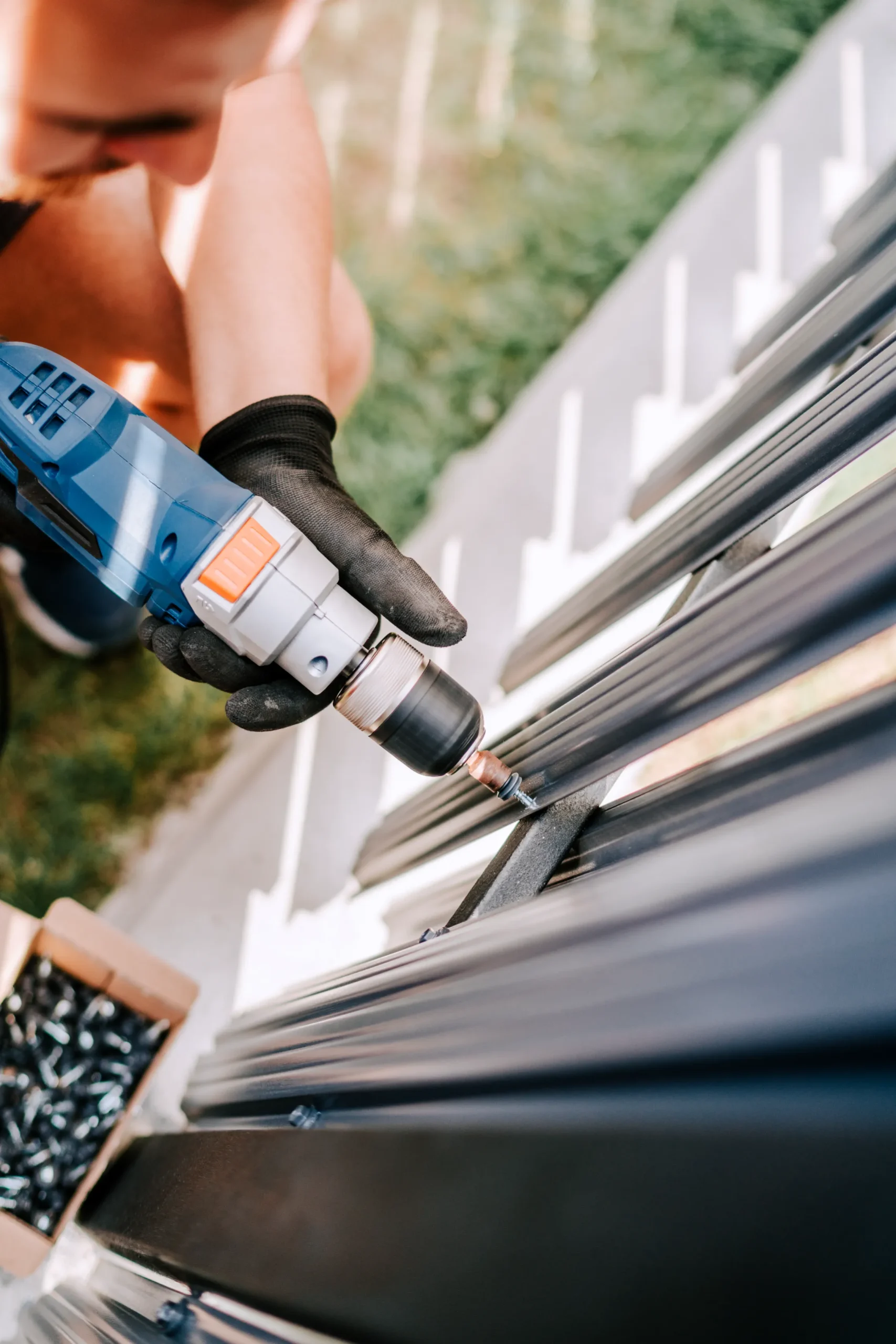 Close-up of a person using a power drill to assemble metal bars. They wear black gloves. A box of screws is nearby. The setting is outdoors.