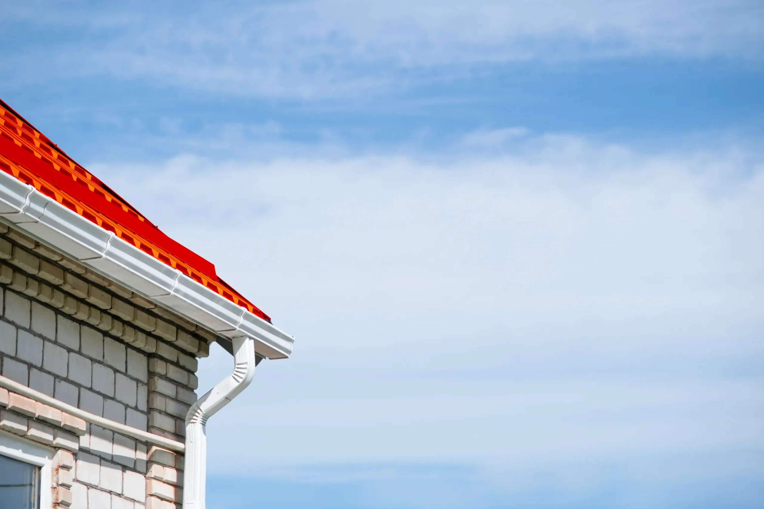 Corner of a house with light-colored brick walls and a bright red roof under a clear blue sky, evoking a sense of calm and simplicity.