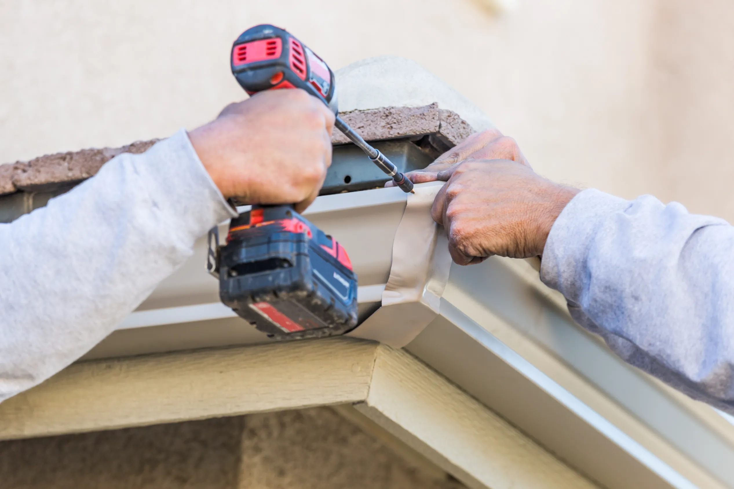 Close-up of two hands installing a white rain gutter on a house. One hand uses a red power drill, while the other holds the gutter in place.
